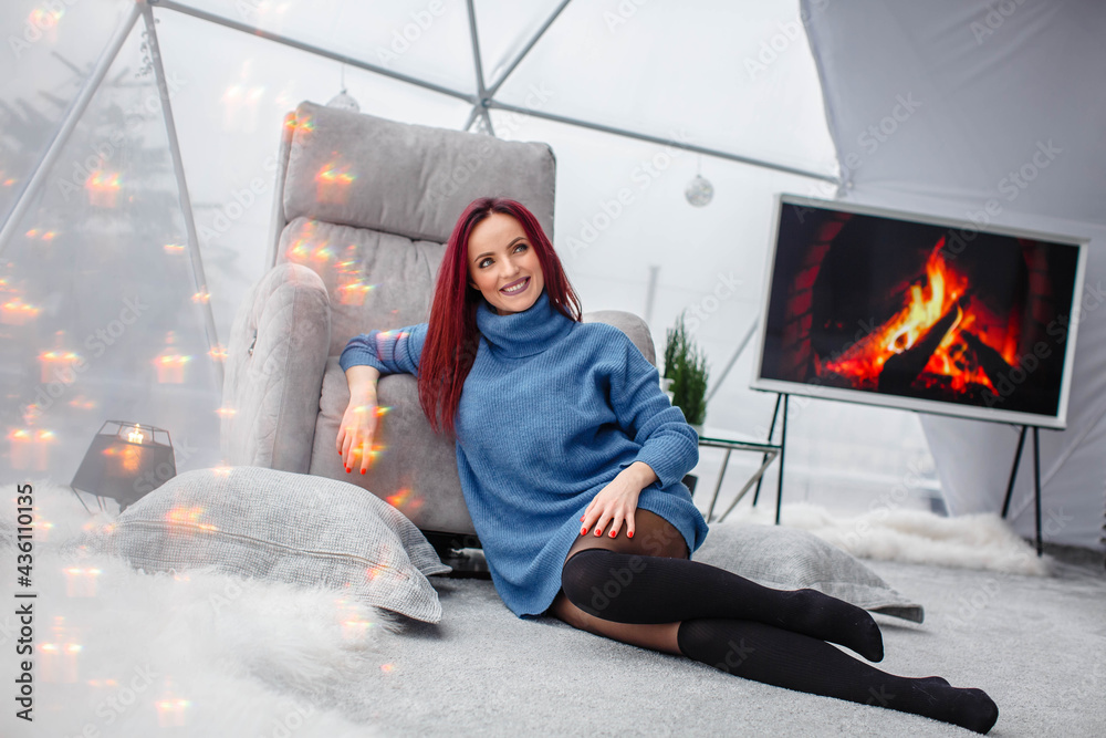 beautiful young girl with red hair and a blue sweater sits near a gray armchair in the living room near the panoramic window and fireplace