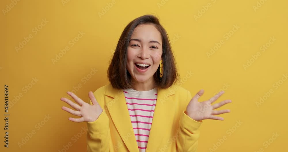 Surprised cheerful Asian woman clasps hands hears awesome news smiles happily exclaims emotionally glad to hear about own success and achievements dressed elegantly isolated on yellow studio wall