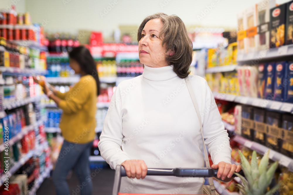 Mature positive woman choosing food products on shelves in grocery shop