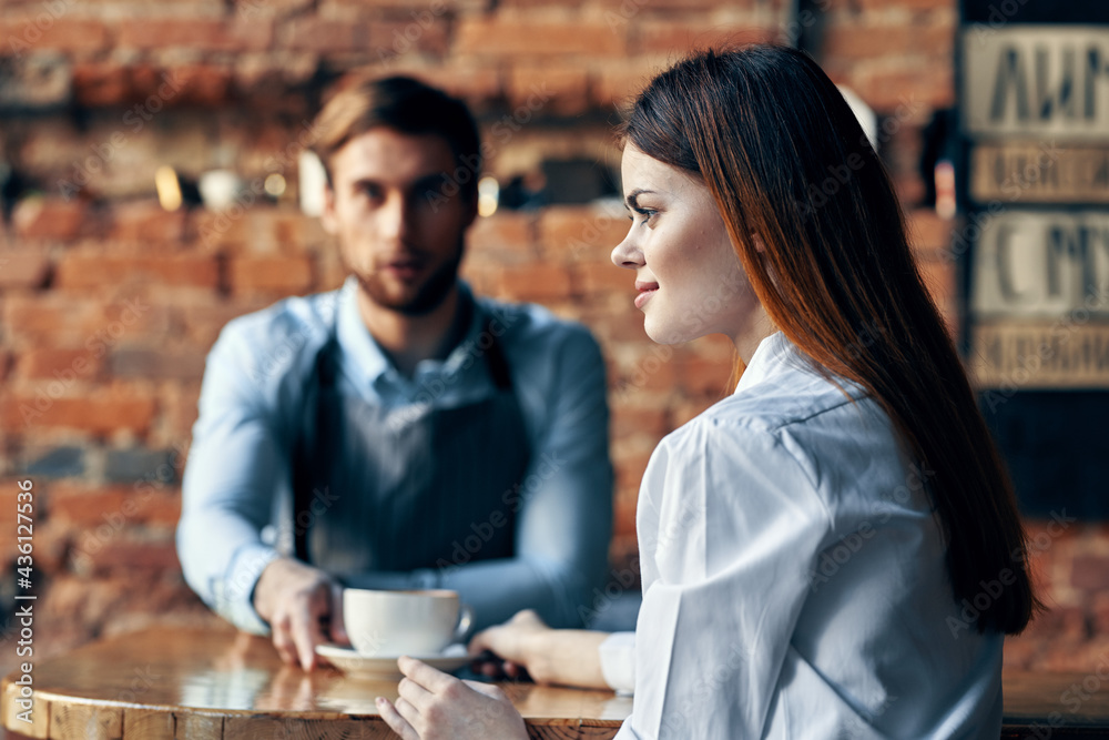 happy woman with a cup of coffee and male bartender in the apron at the table indoors