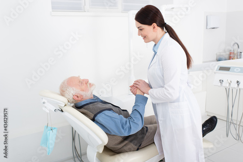 smiling dentist holding hands of senior patient lying in dental chair.