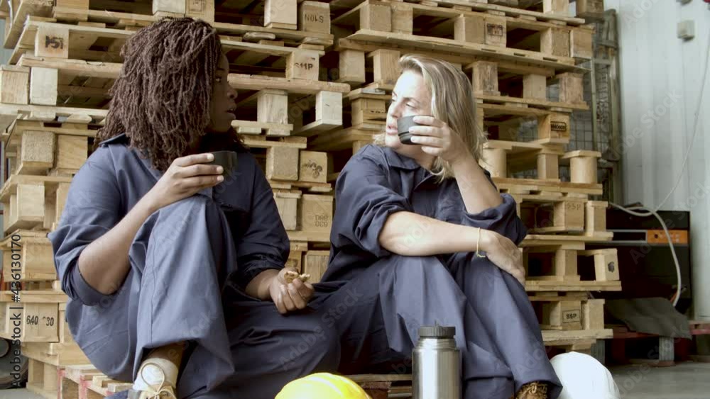 Relaxed women in uniform sitting and drinking coffee. African American woman eating cookies, enjoying time with her colleague, talking, sitting at wooden platform. Warehouse, communication concept. Relaxed women in uniform sitting and drinking coffee. African American woman eating cookies, enjoying time with her colleague, talking, sitting at wooden platform. Warehouse, communication concept.
