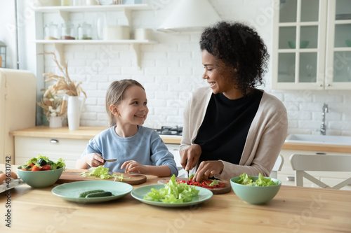 Overjoyed young African American mother and small Caucasian daughter cook healthy tasty salad in kitchen together. Smiling biracial mom and little child have fun preparing food. Adoption concept.