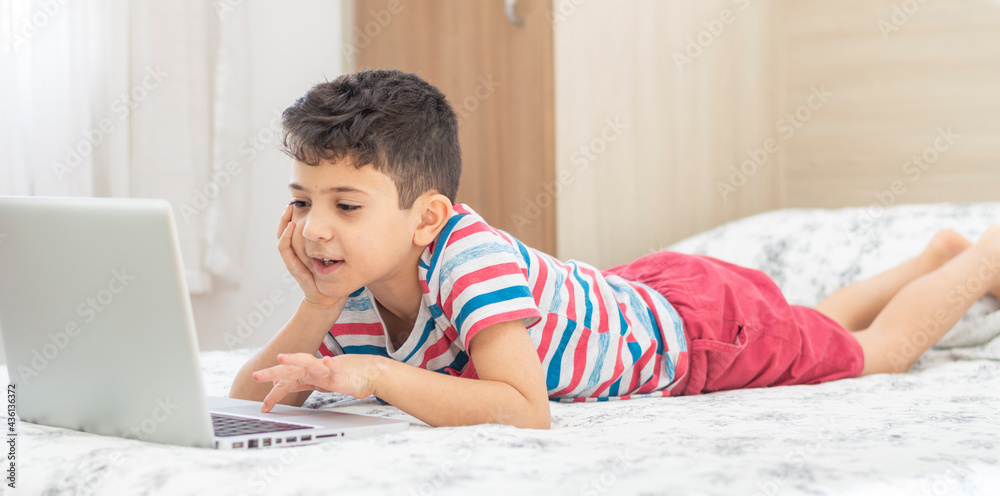 Young boy child lying on bed using a laptop. Online class lesson on video. Distant remote education. New normal school study. Kids at school Young boy child lying on bed using a laptop. Online class lesson on video. Distant remote education. New normal school study. Kids at school