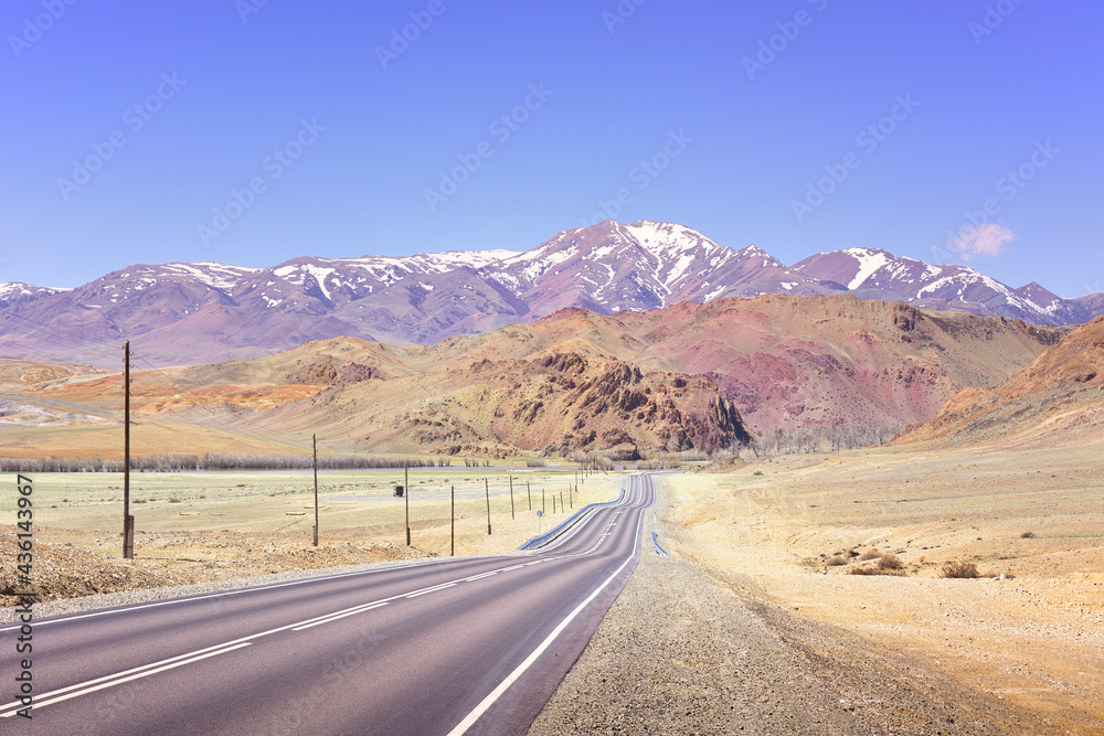 Chuysky tract in the Altai Mountains. Asphalt undulating road in the Chui steppe on the background of snowy mountains under a blue sky. Pure Nature of Siberia, Russia