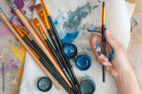 The artist's close-up hand holds a brush against the background of a palette, brushes and paints, is engaged in art, creativity, hobbies, anti-stress or art therapy.