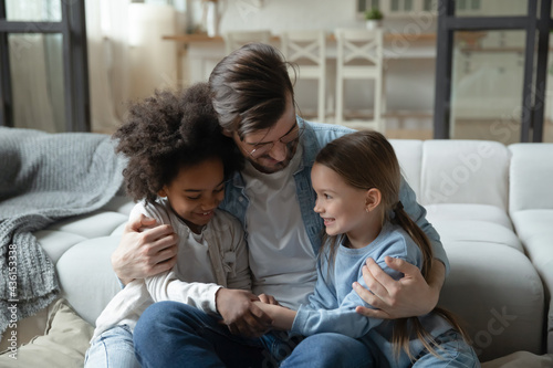 Loving young Caucasian father hug cuddle with two small multiracial daughters, enjoy family weekend at home. Caring dad embrace little multiethnic girls children. Adoption, parenthood concept.