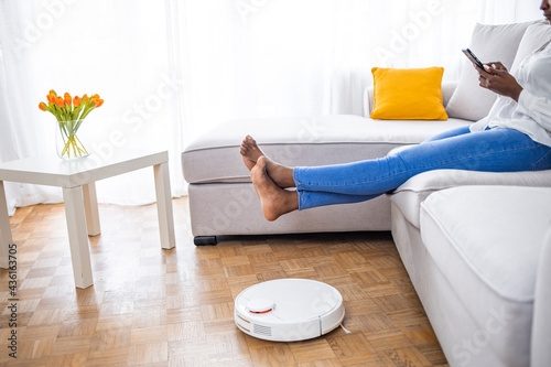 Woman with robotic vacuum cleaner. Robotic vacuum cleaner maintenance. Woman Using Home Robot Cleaning App. Woman with raised legs on sofa and robotic vacuum cleaner on floor in living room