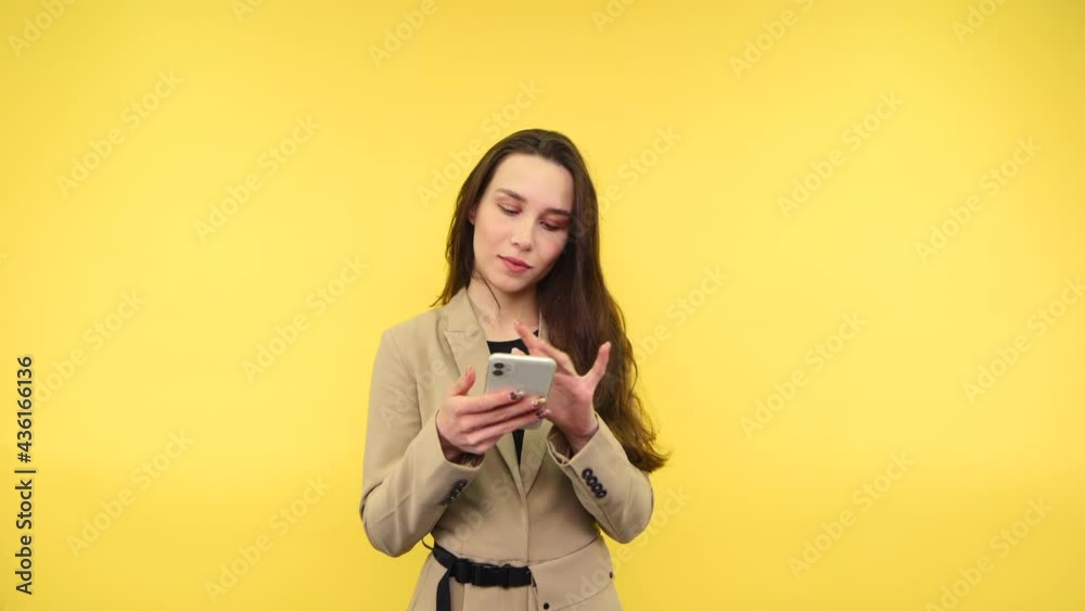 Positive woman in smart casual clothes stands on a yellow background and uses a smartphone with a smile on his face, wearing a beige suit. Positive woman in smart casual clothes stands on a yellow background and uses a smartphone with a smile on his face, wearing a beige suit.