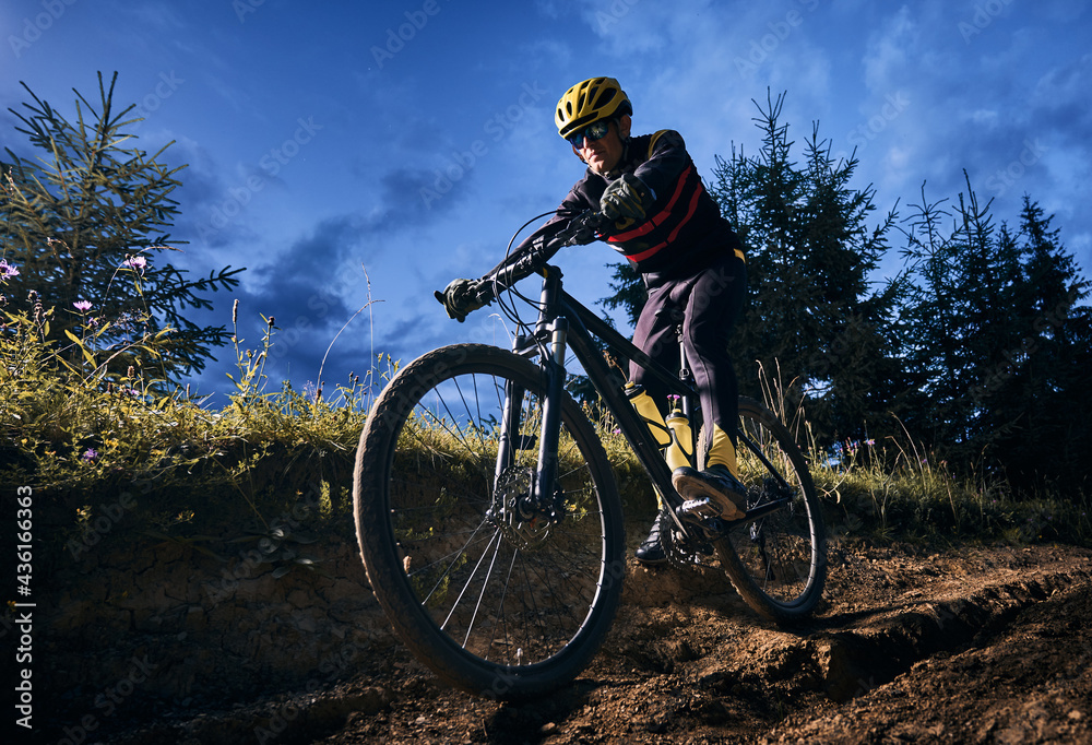 Wide angle view of young man riding bicycle downhill with blue evening sky on background. Bicyclist in cycling suit cycling down grassy hill at night. Concept of sport, biking and active leisure.