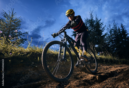 Fototapeta Naklejka Na Ścianę i Meble -  Wide angle view of young man riding bicycle downhill with blue evening sky on background. Bicyclist in cycling suit cycling down grassy hill at night. Concept of sport, biking and active leisure.