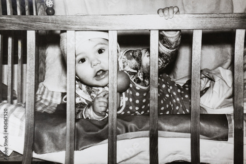 Little baby in bed looking through the wooden rods. Vintage black and white paper photo. Early 1980s. Old surface, soft focus. Transferred property, family archive.