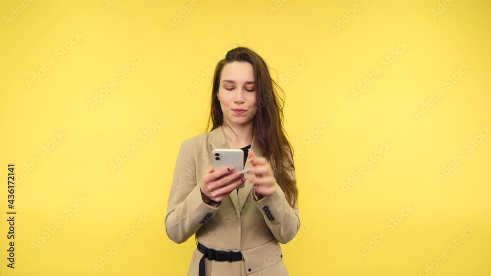 Pensive woman in smart casual clothes stands on a yellow background with a smartphone in her hands and thinks she has found the idea of wearing a beige suit. Pensive woman in smart casual clothes stands on a yellow background with a smartphone in her hands and thinks she has found the idea of wearing a beige suit.