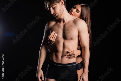 seductive woman in lingerie touching chest of man with shirtless torso and closed eyes on black background