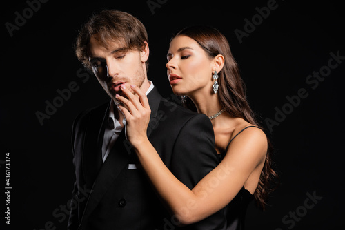 woman in silk dress sensually touching lips of man in black blazer isolated on black