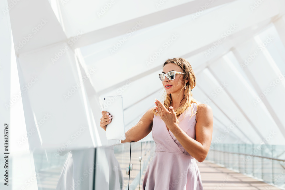 A girl in sunglasses and a summer dress communicates with friends using a tablet, making a video call while walking A girl in sunglasses and a summer dress communicates with friends using a tablet, making a video call while walking