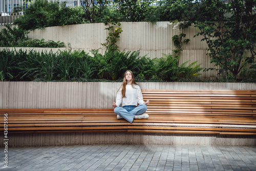 Young attractive woman practicing yoga, sitting in bench, exercise, Lotus pose, working out, wearing casual, white t-shirt, pants, outdoor urban city view.