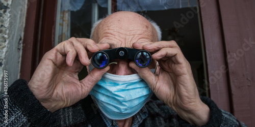 Mature old man wearing medical face mask stands on balcony with binoculars