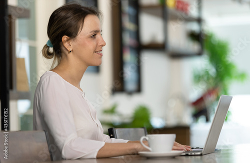 Beautiful young Caucasian woman freelancer in white long-sleeved shirt sitting at ease in cafe on afternoon and typing on laptop for working online business