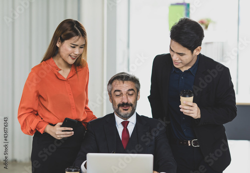 Senior Caucasian businessman sitting in office while Asian colleagues of woman in orange shirt and man in black suit standing behind smiling looking at laptop together as happy for job success