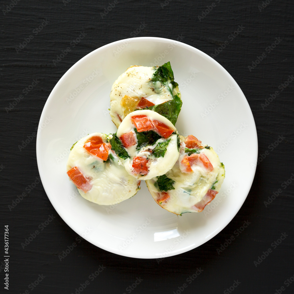 Homemade Egg White Breakfast Cups with Spinach and Tomato on a white plate on a black background, top view. Flat lay, overhead, from above. Homemade Egg White Breakfast Cups with Spinach and Tomato on a white plate on a black background, top view. Flat lay, overhead, from above.