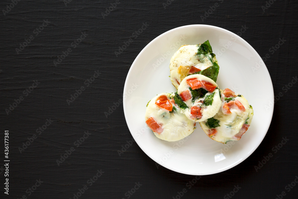 Homemade Egg White Breakfast Cups with Spinach and Tomato on a white plate on a black background, top view. Homemade Egg White Breakfast Cups with Spinach and Tomato on a white plate on a black background, top view.