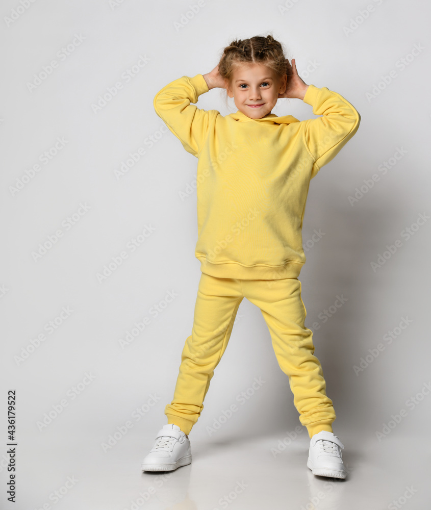Full length portrait of a little girl dressed in a bright yellow sports suit. Cute child posing on a white background with arms up. Concept of active childhood.