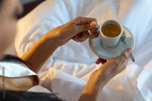 A young female, wearing a skin-colored nightdress and holding a cup of coffee in her hands in bed on a white blanket. Concept of a relaxed morning in bed.