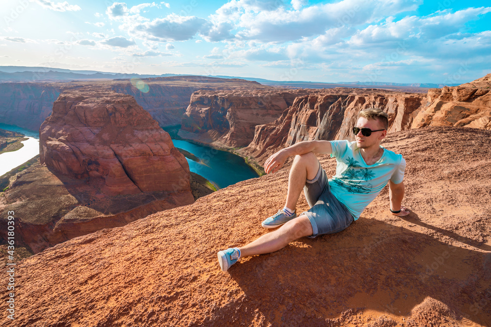 Panoramic view of a young woman sitting on the edge of a cliff facing the Horseshoe Bend, a horseshoe meander of the Colorado River in the town of Page, Arizona