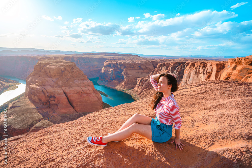 A beautiful young woman sits on the edge of a cliff overlooking Horseshoe Bend, the horseshoe meander of the Colorado River in the city of Page, Arizona