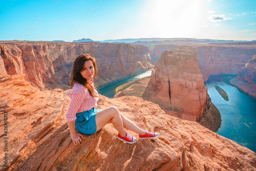 A beautiful young woman sits on the edge of a cliff overlooking Horseshoe Bend, the horseshoe meander of the Colorado River in the city of Page, Arizona