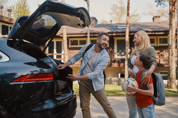 Happy young family with little packing stuff into the car and smiling while standing near house