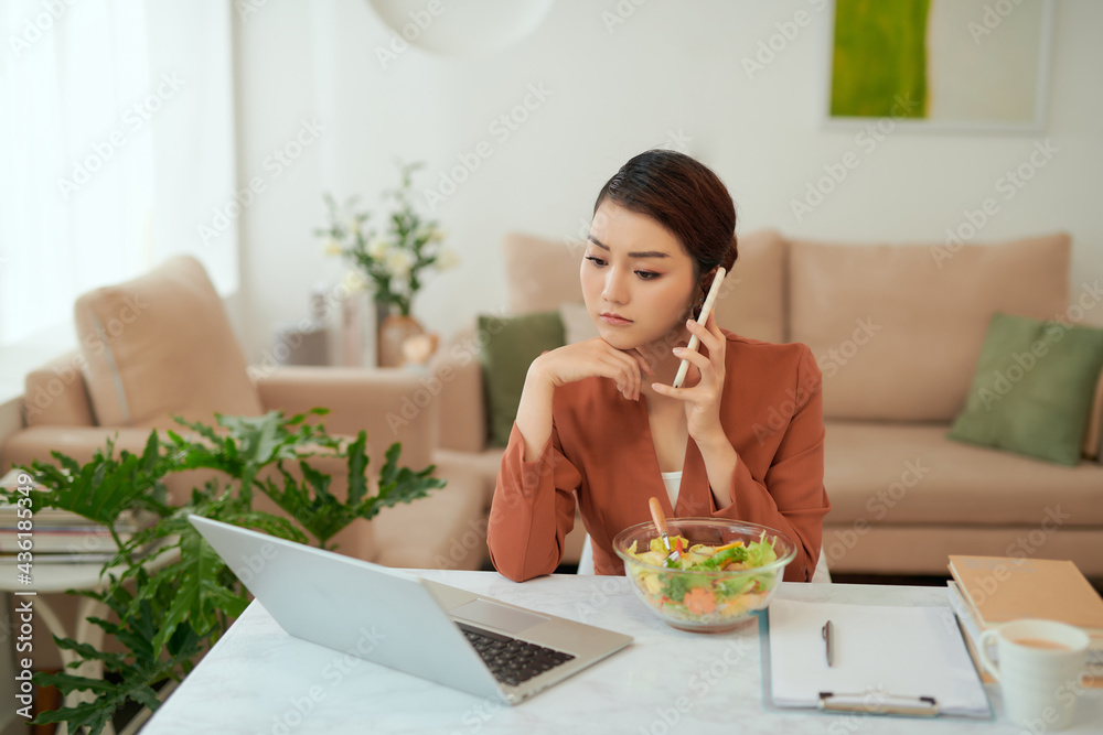 Pretty young woman eating healthy salad in office while talking on mobile phone