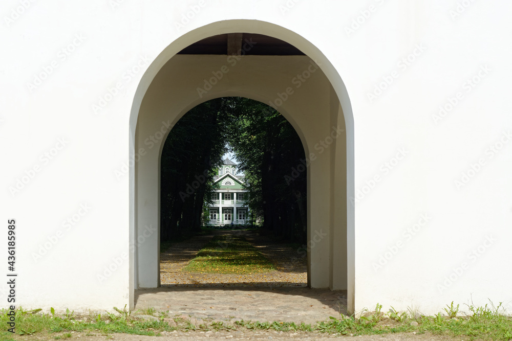 View of the wooden house through the arch of the observation gazebo in the Petrovskoye estate in the Pushkinskie Gory. View of the wooden house through the arch of the observation gazebo in the Petrovskoye estate in the Pushkinskie Gory.