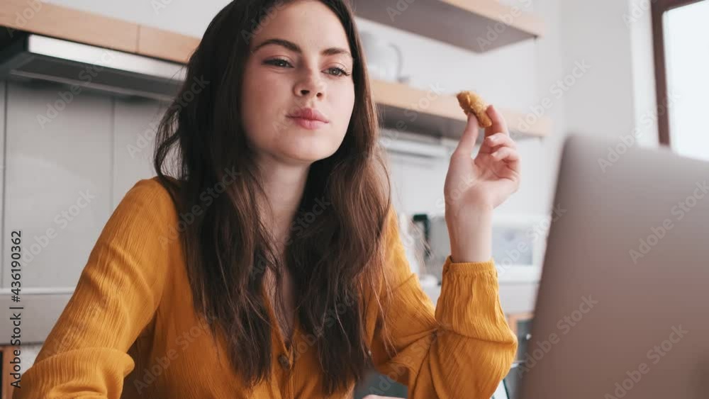 A smiling woman eating the cookies and looking at the laptop in kitchen A smiling woman eating the cookies and looking at the laptop in kitchen