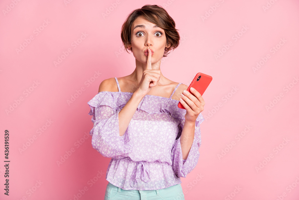 Photo portrait of girl covering mouth with finger holding phone in one hand isolated on pastel pink colored background