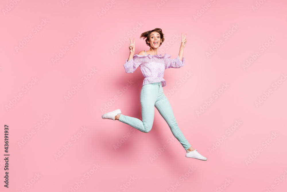Full size photo of young happy charming excited positive girl jumping showing v-sign isolated on pink color background