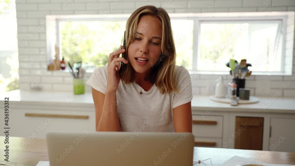 Caucasian female working in kitchen chatting to friends on cellular device typing on laptop Caucasian female working in kitchen chatting to friends on cellular device typing on laptop