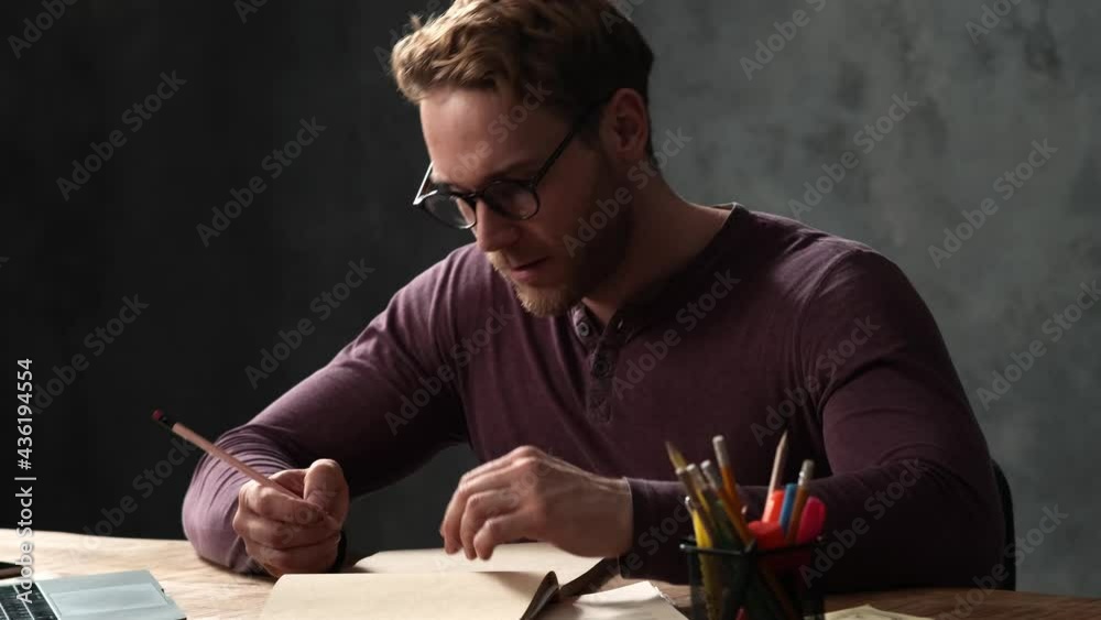 A smiling man with glasses looking at a laptop and then thinking what to write in a notebook while sitting at a table with a laptop in an office with a gray wall