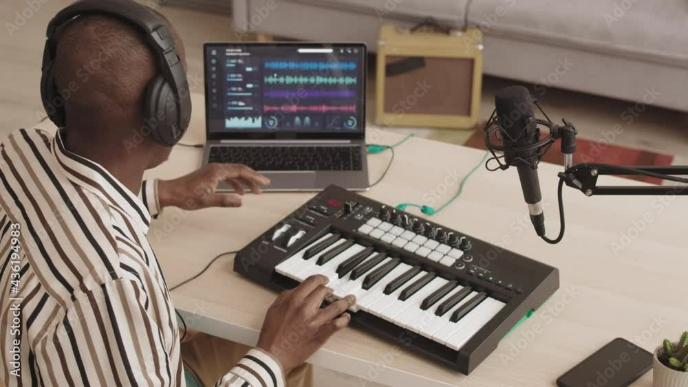 High angle view of bald African-American male artist sitting in home music production studio, singing while playing modern midi keyboard synthesizer