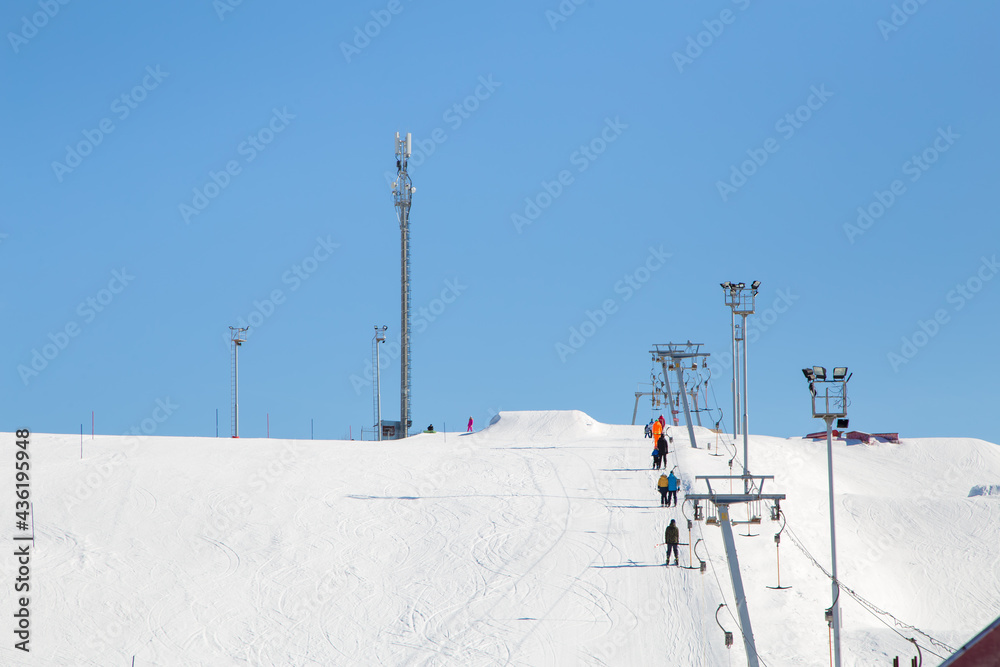 Ski resort, gentle snow slope with people on the lift going to the top. Mountain slope for skiing and snowboarding. Against the background of the blue sky. The summit and ski tours on a sunny day.