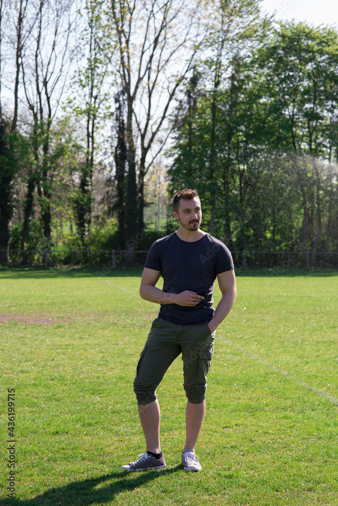 Obraz premium Hübsche jünger Mann mit schwarzen Haaren, Bart, steht auf dem grünen Rasen Er schaut zur Seite und genießt das schöne Sommerwetter, Natur und das Leben. Er sieht toll aus. 