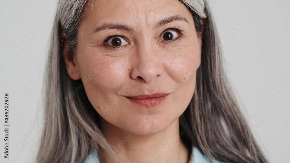 A close-up view of a smiling elder woman is showing silence gesture to the camera standing isolated over a grey wall in the studio A close-up view of a smiling elder woman is showing silence gesture to the camera standing isolated over a grey wall in the studio
