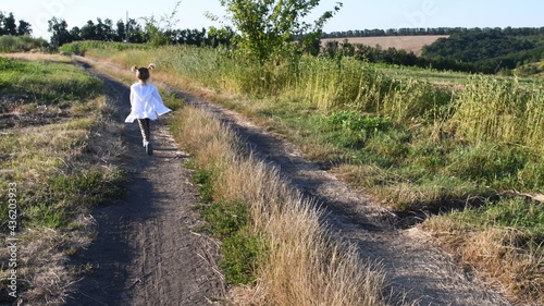 Defocused silhouette of little girl running away by dirt road at rural landscape in summer. White clothes fly in wind. Serenity of childhood