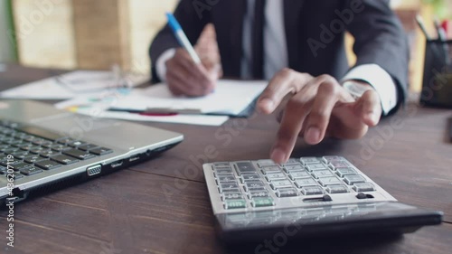 Black man office worker in a suit counts on calculator and records the results with a pen in paper tablet while sitting at desk with laptop the office. Concept of accounting and savings. Bottom view photo