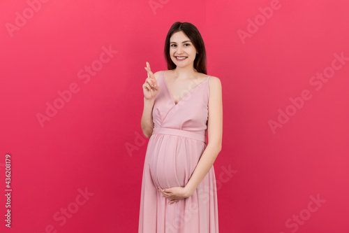 Cheerful young pregnant woman holding fingers crossed, waiting pregnancy isolated on colored background in studio. lifestyle pregnancy concept © sosiukin