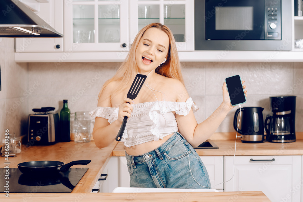  Woman singing and dancing in the kitchen with a smartphone, music.A girl in headphones with a phone stands in the kitchen and prepares food behind an induction stove.