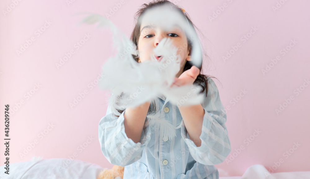 Fototapeta premium Closeup portrait of a cute little girl in pajama blowing white feathers while playing in the bed, on pink background. A kid playing games in the morning.