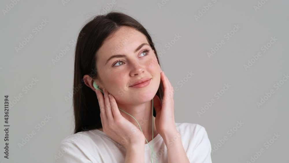 A beautiful woman wearing basic T-shirt is listening the music wearing wired earphones standing isolated over grey background in the studio A beautiful woman wearing basic T-shirt is listening the music wearing wired earphones standing isolated over grey background in the studio