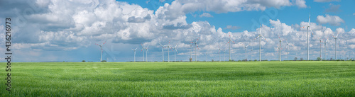 Panoramic view over beautiful farm landscape with green wheat field and wind turbines to produce green energy in Germany, Spring, at blue dramatic sky with clouds and sunny day.